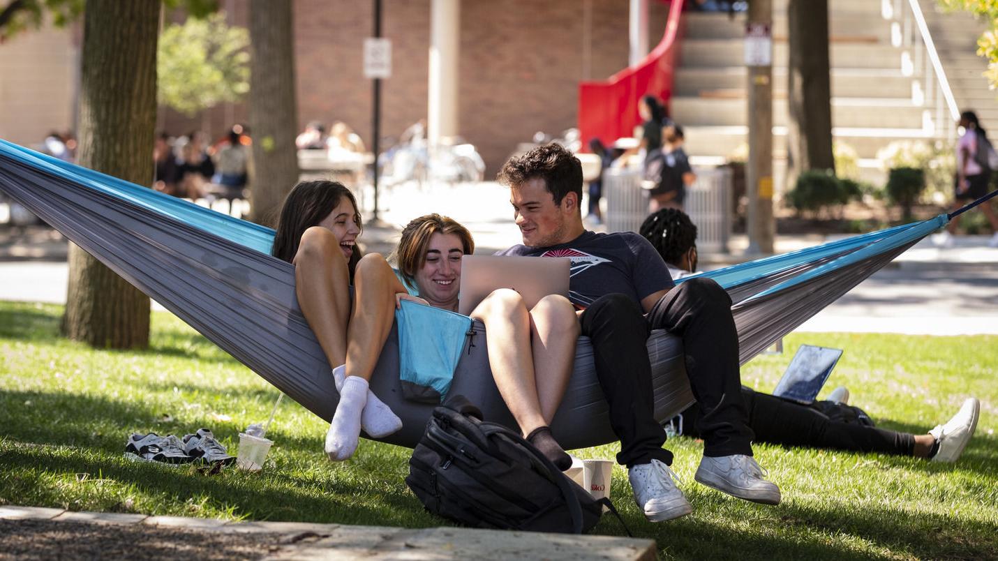 Three friends sit on a hammock on campus.